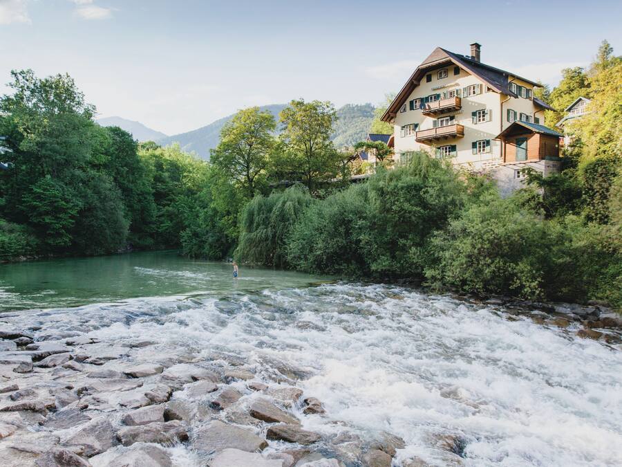 A riverside hotel with a view of a flowing river, surrounded by lush greenery and mountains in the background.