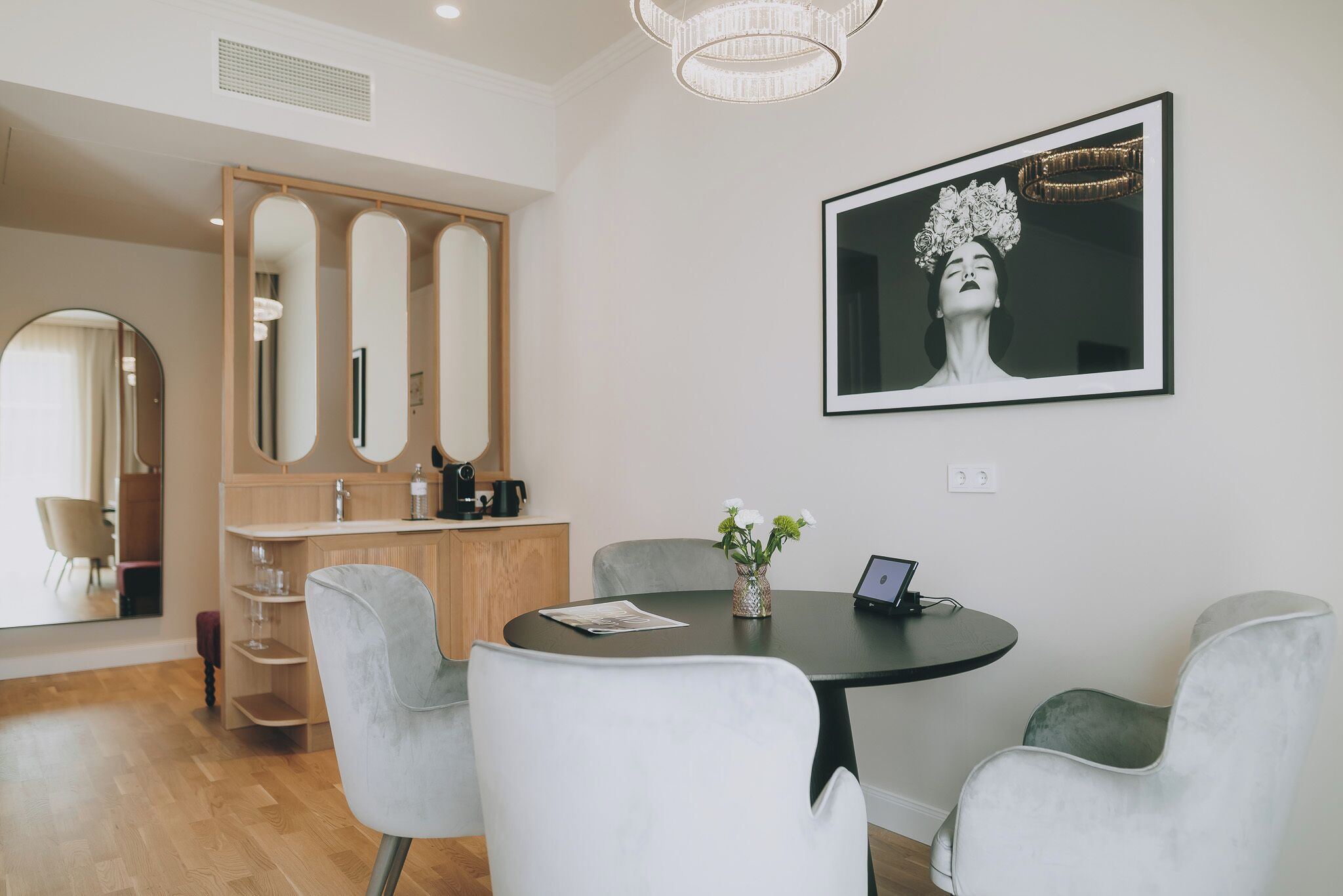 A modern dining area with a round table, white chairs, and a vase with flowers. There is a framed artwork on the wall and a wooden cabinet with mirrors.