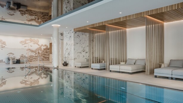 A modern indoor pool area with a glass barrier, seating along the pool, and a decorative wall with a tree mural. - Grand Elisabeth (Bad Ischl)