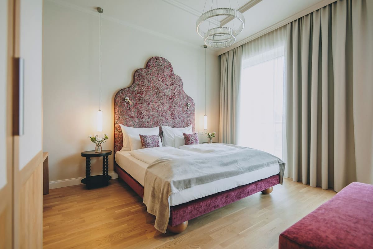 A neatly arranged bedroom with a large bed, a bedside table with a lamp, and a view of the outside through a window with curtains.
