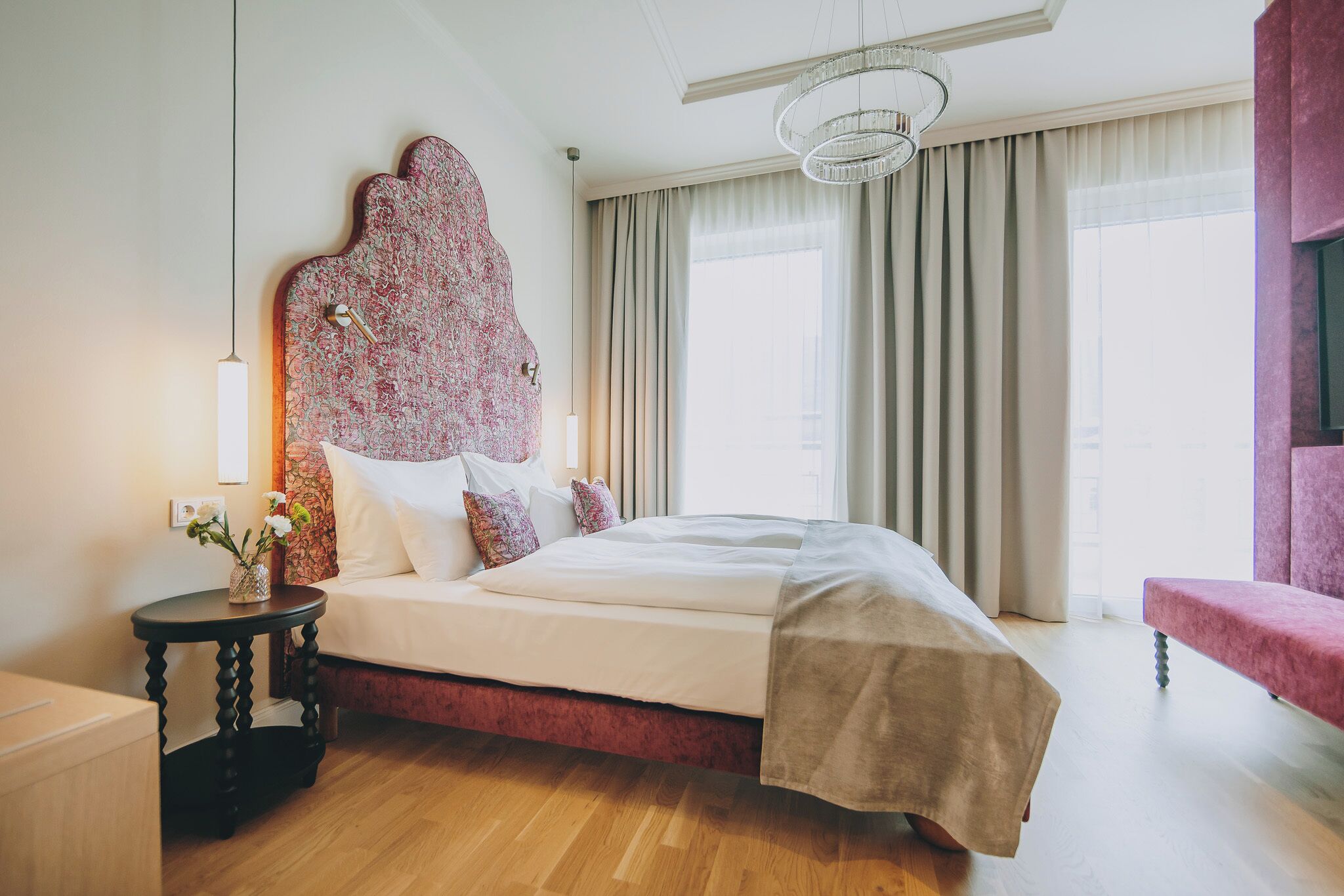 Una camera d'albergo moderna con un letto grande, una testiera decorativa, un tavolino rotondo e vista sulla città attraverso ampie finestre.