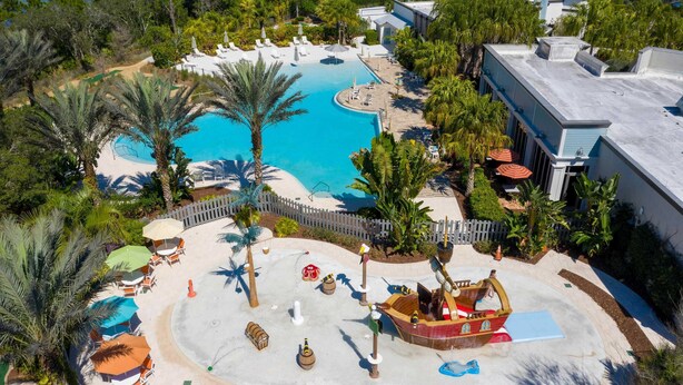 An aerial view of a resort with a large pool, sun loungers, palm trees, and a pirate ship play area.
