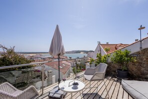 A balcony with wicker chairs, a table with wine glasses and a bowl of olives, a potted plant, and a view of the sea and buildings. - J ia - No Centro Hist Rico da Vila (Alcobaça)