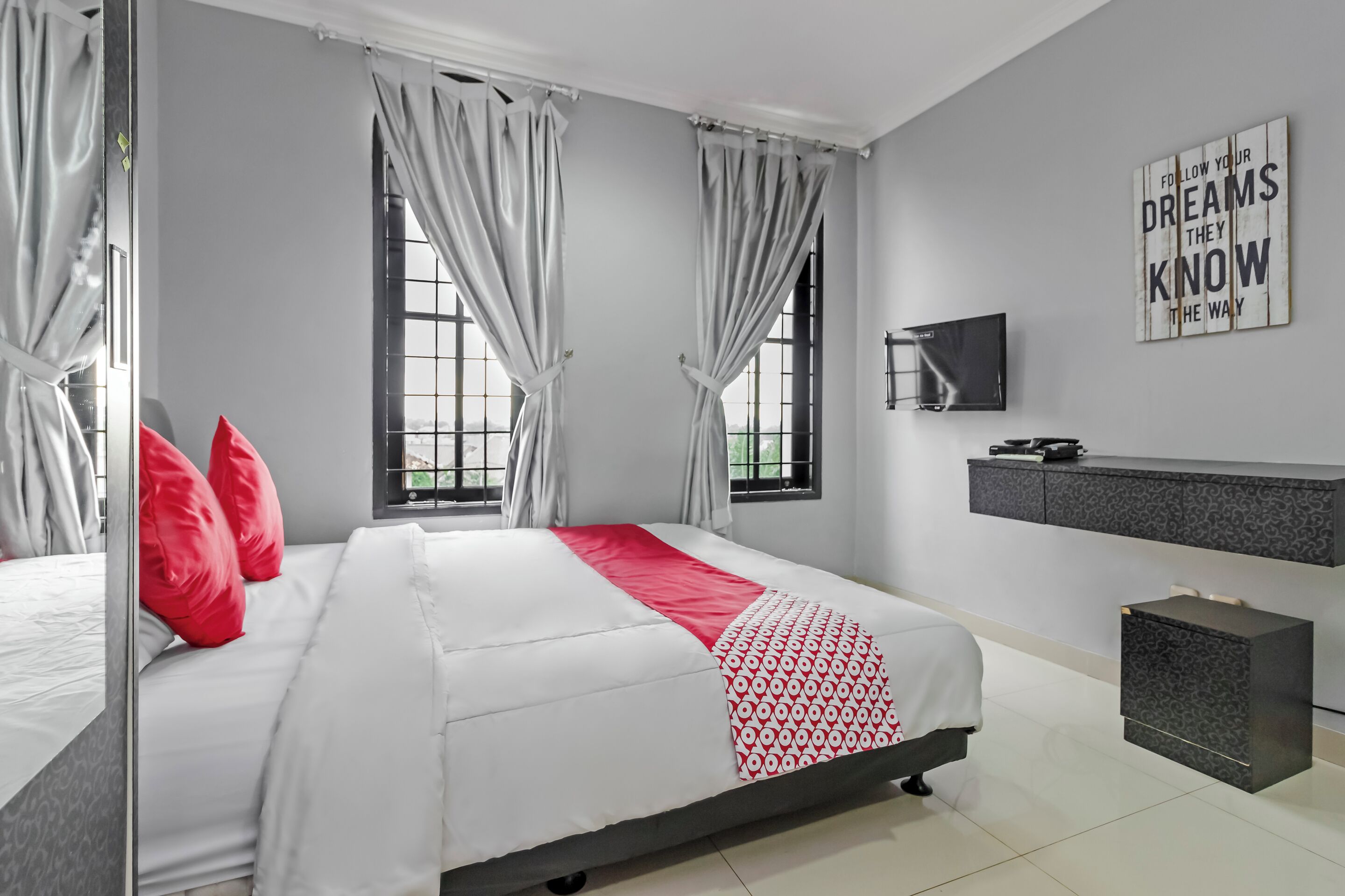 A modern bedroom with a bed, red pillows, a patterned throw, a TV, and a grey console.