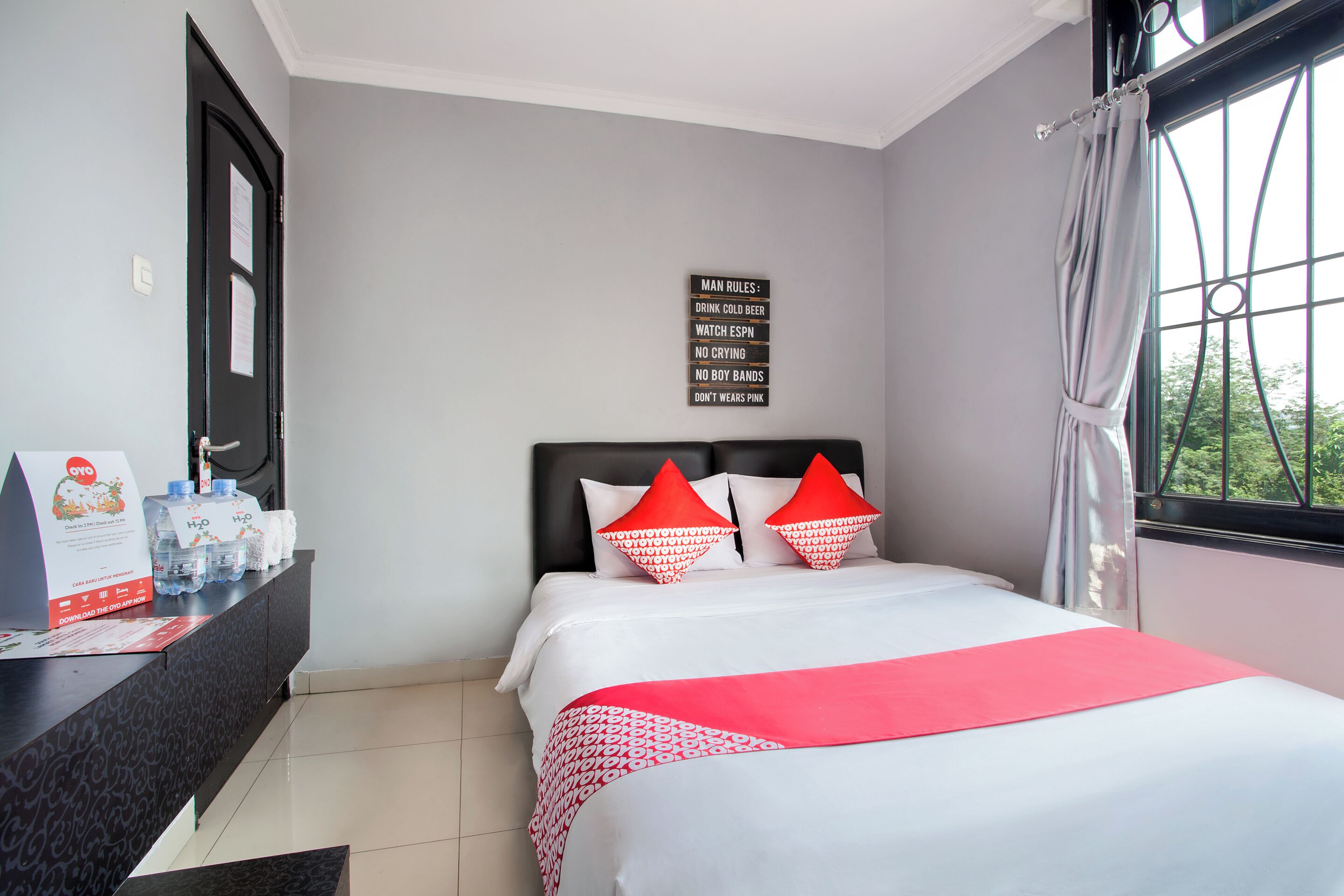Une chambre d’hôtel avec un lit, une table de chevet avec une bouteille d’eau, un panneau indiquant les règles, et une fenêtre avec des rideaux.