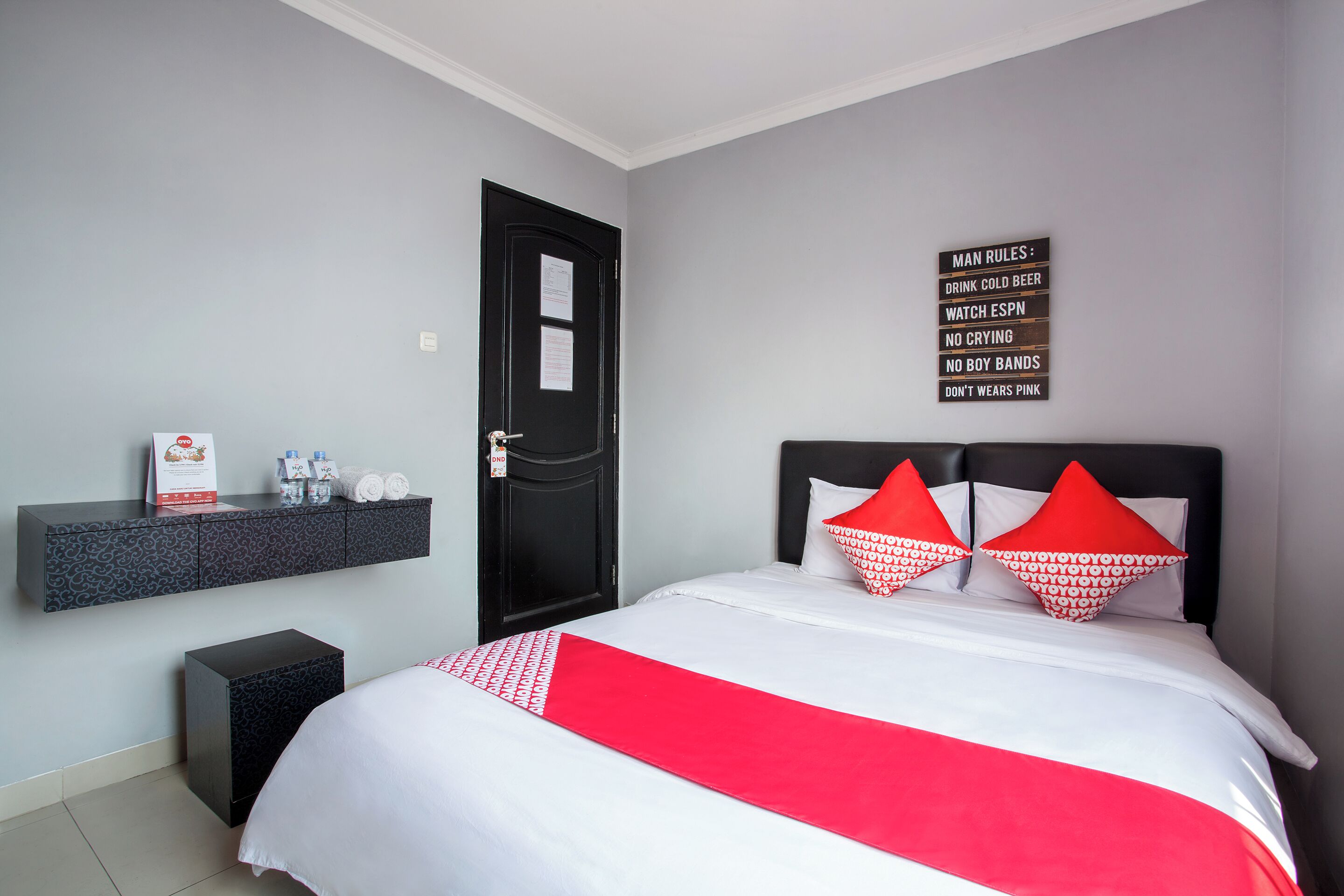 A hotel room with a bed, a black door, a wall-mounted shelf with toiletries, and a sign listing man rules.