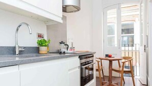 A modern kitchen with white cabinets, a stainless steel sink, and a built-in oven. There is a small wooden table with a chair and a potted plant on the counter.