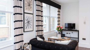 A modern living room with a black sofa, a small side table with a lamp, a flat-screen TV on a black stand, and a window with striped curtains.