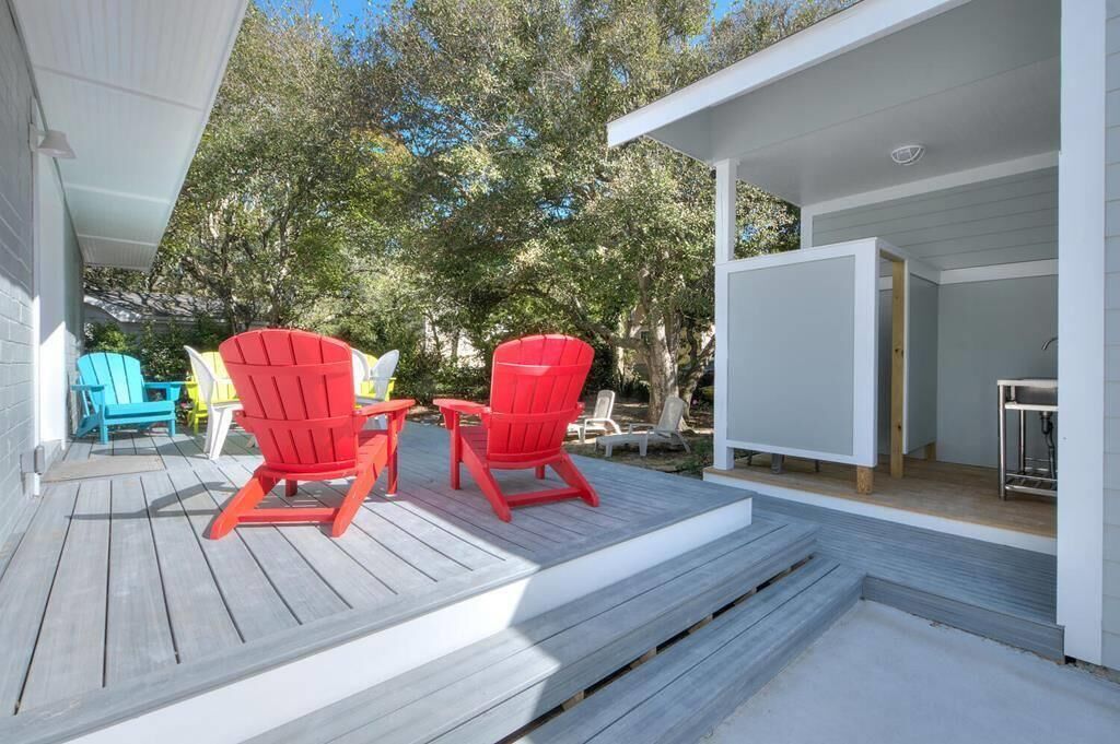 A wooden deck with red Adirondack chairs and a small table, a covered area with a table and chairs, and a view of trees and a building in the background.