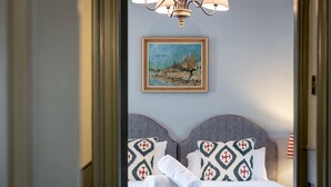 A neatly made bed with patterned pillows and a rolled towel, a painting of a coastal scene on the wall, and a chandelier above.
