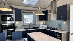 A modern kitchen with a built-in oven, a sink, and a stove. There is a dining area with blue chairs and a table. - Lodge No 20 | Hot Tub |  Stewart's Resort Cameron (Cameron)