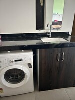Μια μικρή, λειτουργική κουζίνα με εντοιχισμένο πλυντήριο ρούχων, νεροχύτη και σκούρα ξύλινα ντουλάπια.