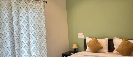 A hotel room with a bed, a ceiling fan, an air conditioner, a nightstand, a lamp, and a patterned curtain.