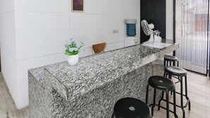 A kitchen area with a granite countertop, a small potted plant, a water dispenser, and a fan. There are three black stools and a chair. - Hotel O Pondok Indah Near Gandaria City Formerly Odori Residence (Jakarta)