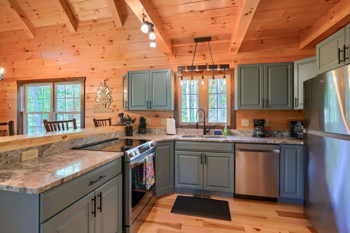 A kitchen with wooden ceiling beams, a stainless steel refrigerator, and a window with a view of trees.