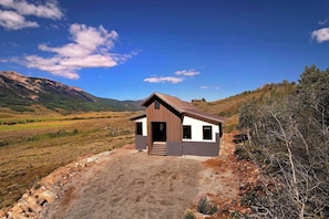 A single-story cabin with a front door, a small porch, and a dirt driveway leading to it.