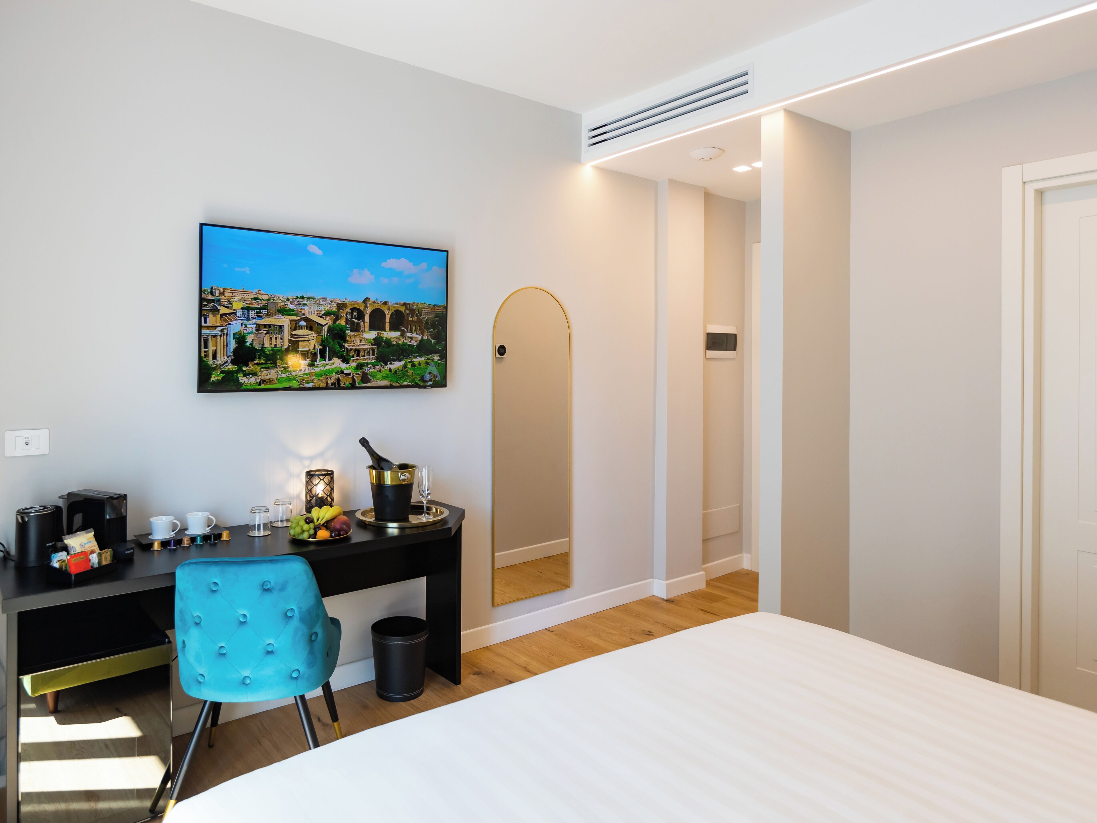 A modern hotel room with a bed, a desk with a computer, a chair, a trash bin, and a wall-mounted TV displaying a cityscape.