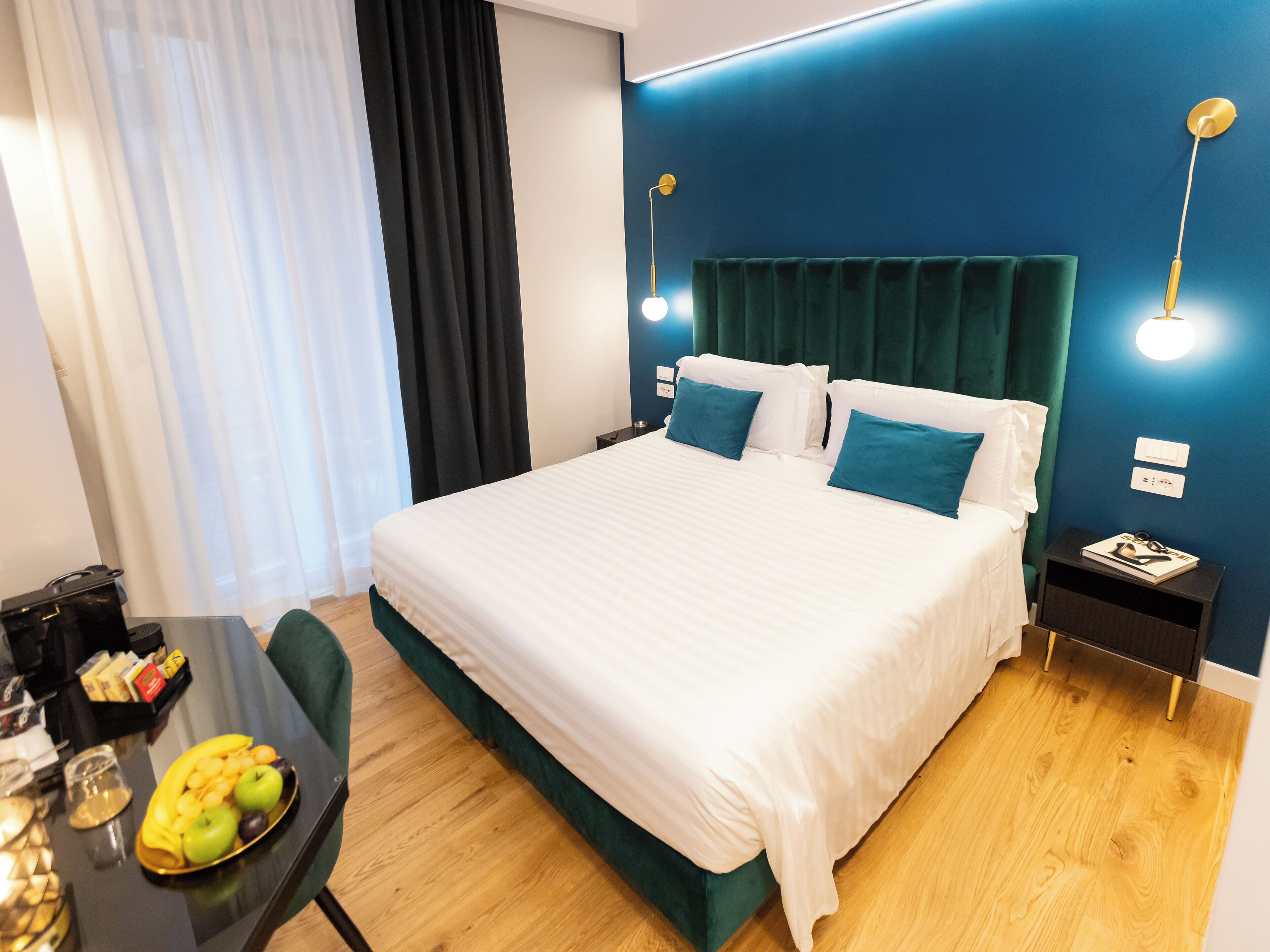 A neatly made bed with white linens and teal pillows, a small bedside table with a lamp, and a fruit bowl on a wooden table.