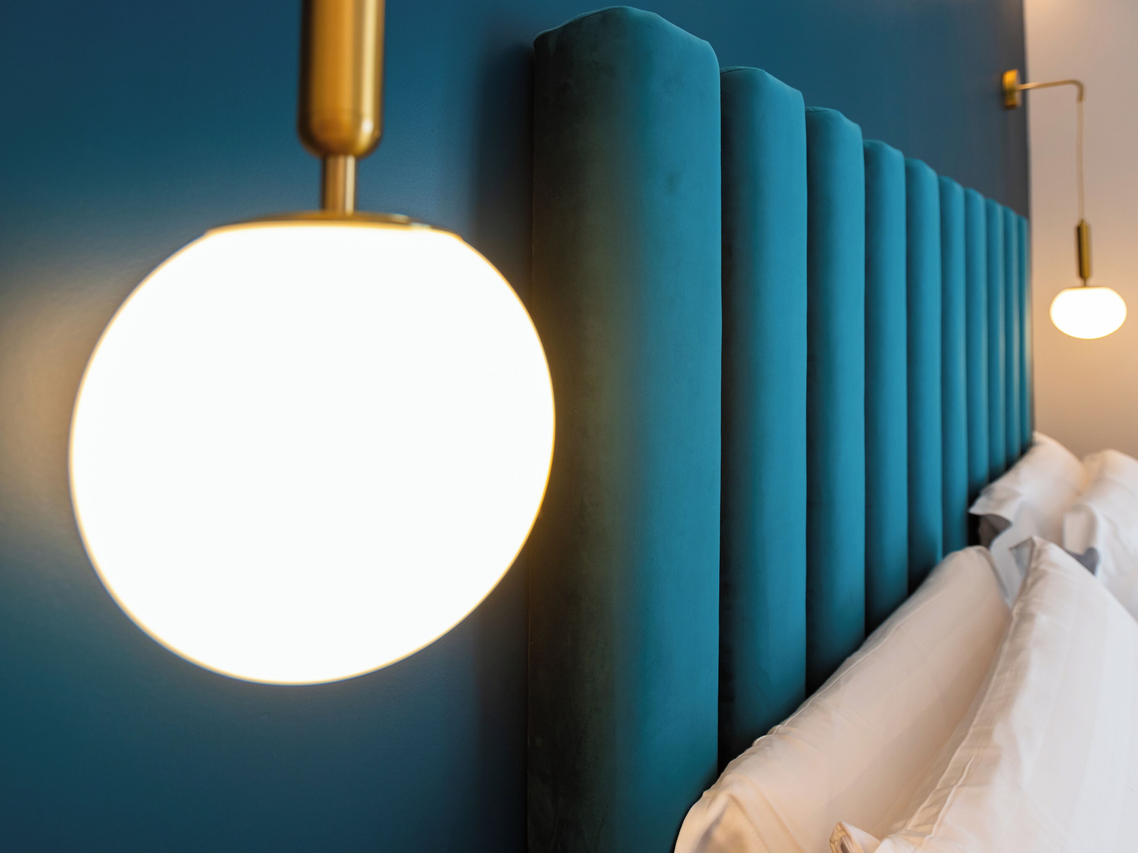 Une chambre moderne avec une tête de lit de couleur turquoise, un luminaire rond fixé au mur et un couvre-lit blanc.