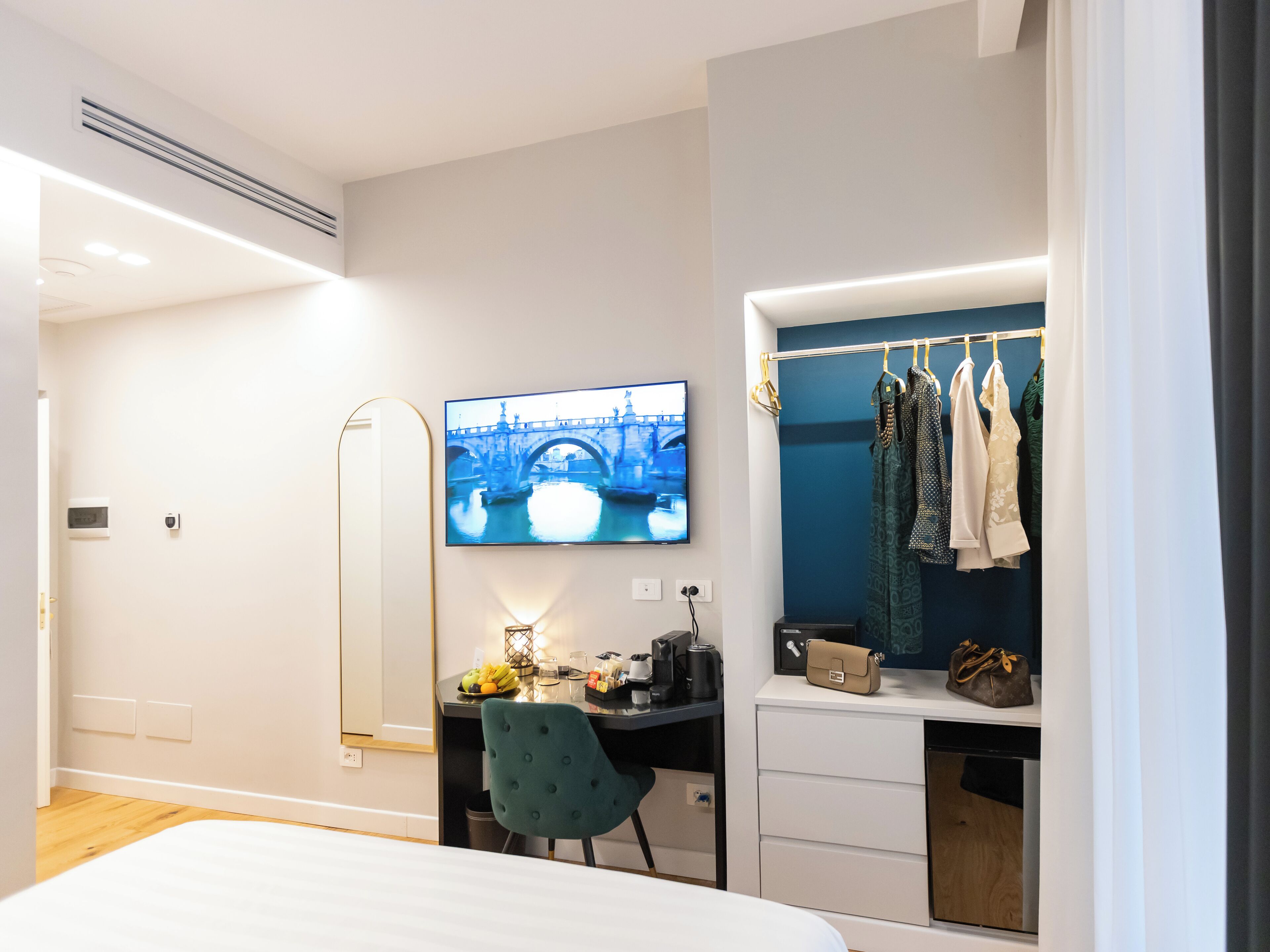 Una habitación de hotel moderna con una cama, un escritorio con una computadora, un televisor mostrando un paisaje urbano, un armario con ropa y una pequeña mesa con una lámpara y un frutero.