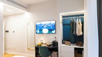 Una habitación de hotel moderna con una cama, un escritorio con una computadora, un televisor mostrando un paisaje urbano, un armario con ropa y una pequeña mesa con una lámpara y un frutero.