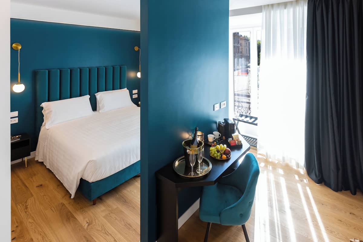 A modern hotel room with a teal-colored headboard, a bed with white linens, a desk with a tray of fruits and a bottle of champagne, and a view of a cityscape through large windows.