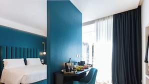 Une chambre d’hôtel moderne avec un mur d’accent de couleur turquoise, un lit avec du linge de maison blanc, un bureau avec une bouteille de champagne, des verres et des fruits, et une grande fenêtre avec des rideaux.