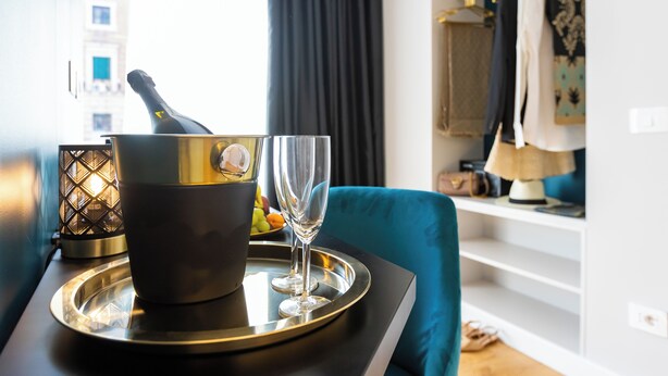 Une pièce avec une table à manger dressée, agrémentée d’une bouteille de champagne, de verres et d’un panier de fruits. On aperçoit une chaise bleue et une armoire à l’arrière-plan.