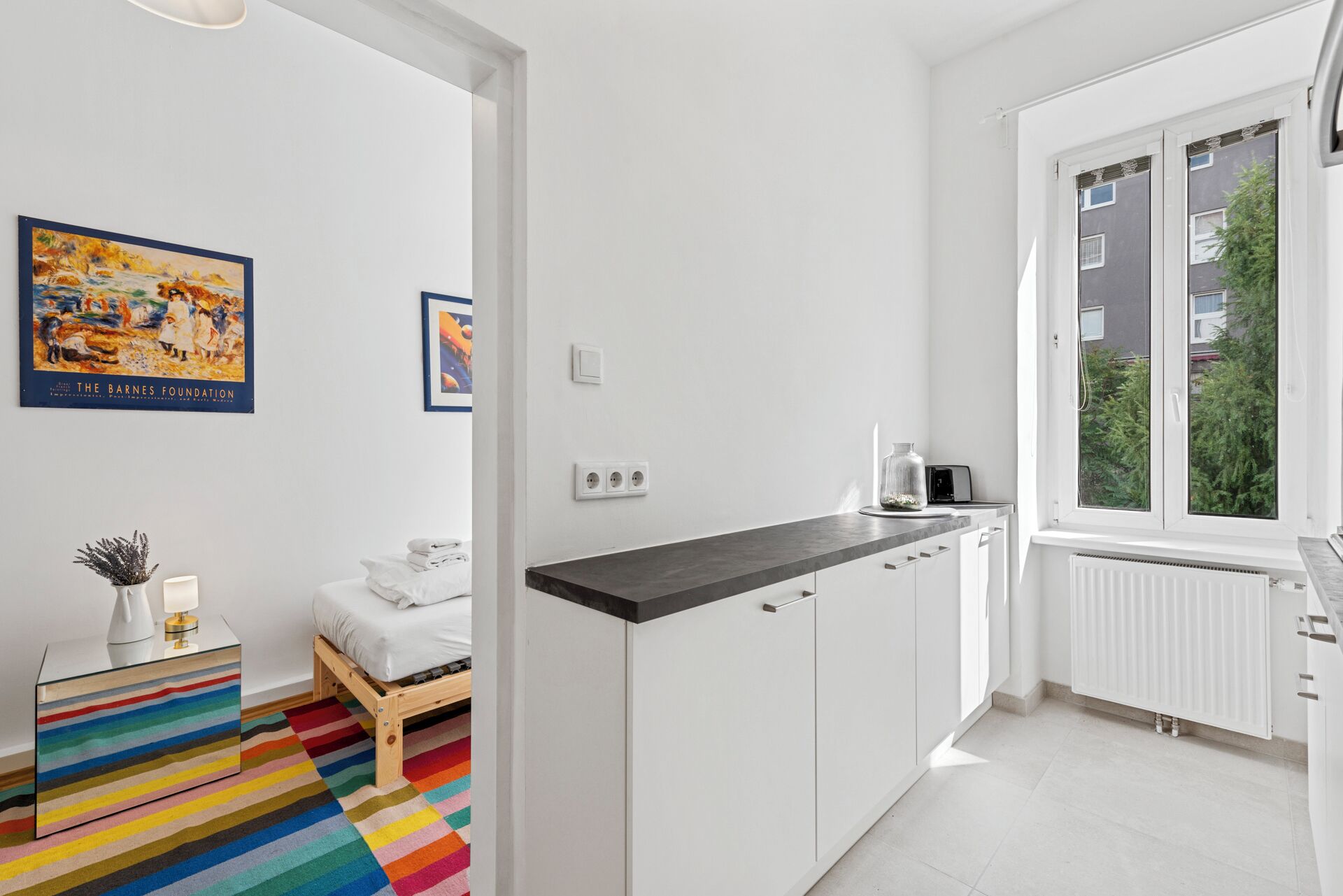 Een moderne keuken met witte kasten, een zwart aanrecht en een raam met uitzicht op groen. In de aangrenzende kamer staat een bed met een vloerkleed met regenboogstrepen.