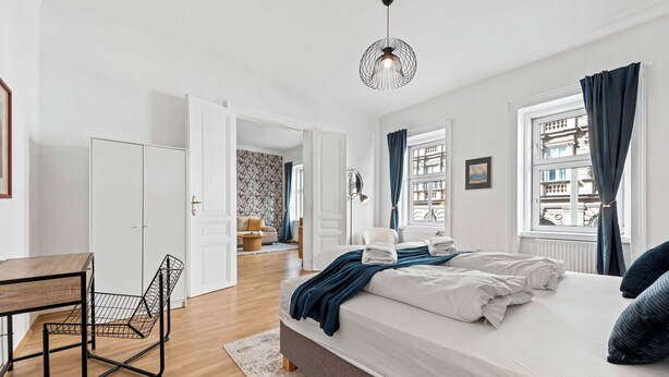 Un dormitorio moderno con una cama grande, un escritorio y una silla. Hay una ventana con cortinas y vistas a edificios.