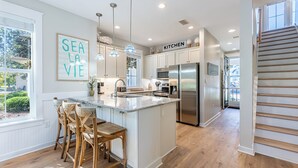 A modern kitchen with a central island, wooden chairs, pendant lights, and a stainless steel refrigerator. - Two Golf Carts ~ Lake & Golf Course Views (Miramar Beach)