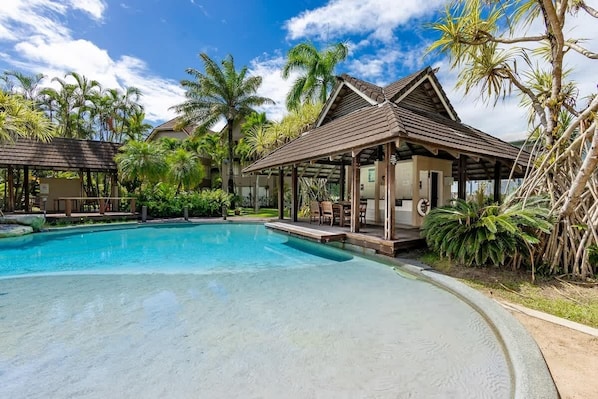 A swimming pool with a wooden deck and lounge chairs, surrounded by palm trees and a clear sky.
