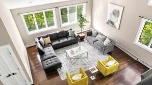 Un salon spacieux comprenant un canapé d’angle, deux fauteuils, une table basse et une grande fenêtre donnant sur la verdure.
