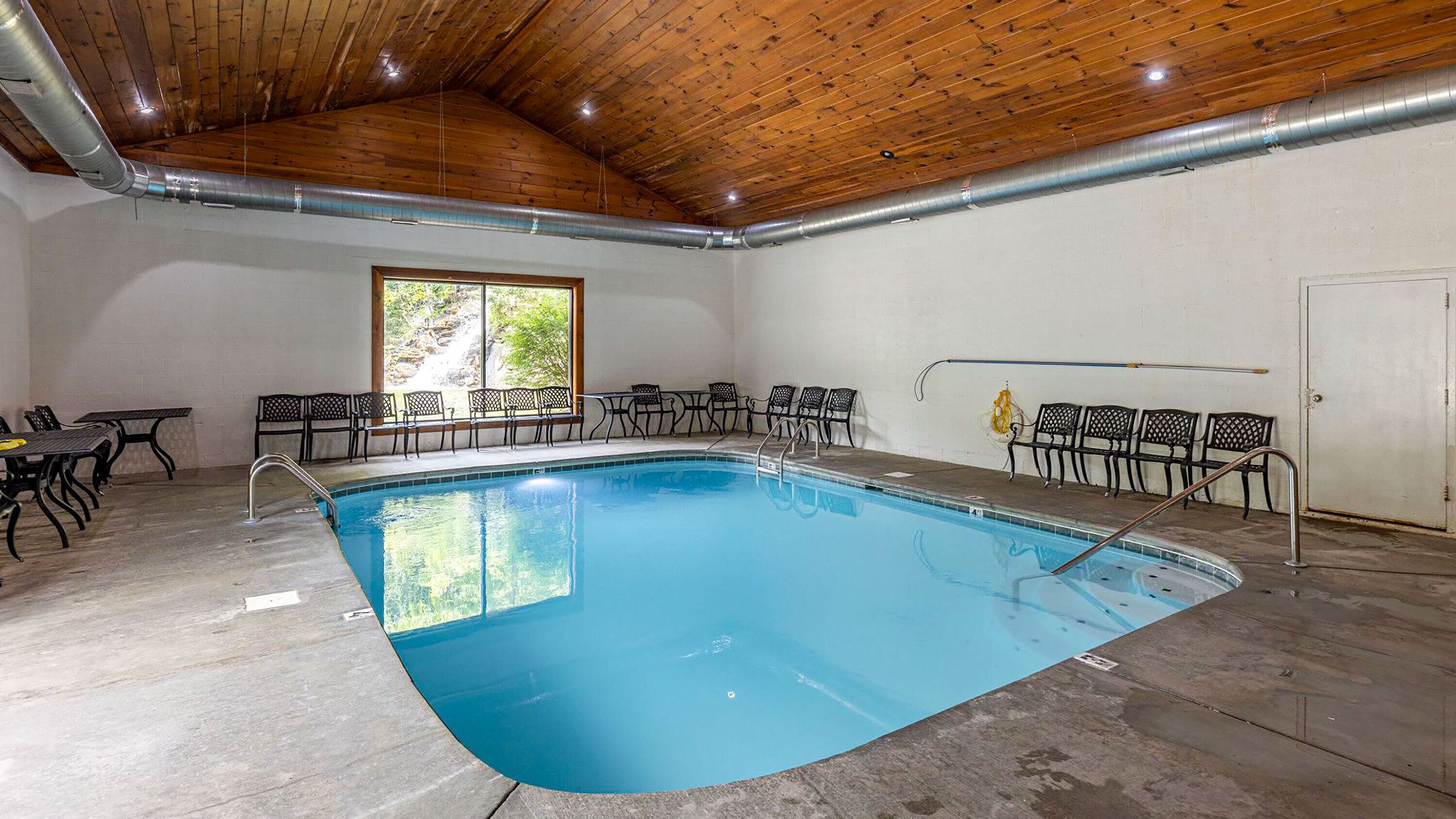 An indoor swimming pool with a wooden ceiling, metal benches, and a view of greenery outside.