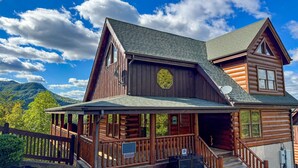 A log cabin with a porch, a satellite dish, and a mountain backdrop. - Evergreen Oasis - 6BR, Theater Room & 2 Hot Tubs!! (Sevierville)