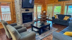 A cozy living room with a stone fireplace, comfortable seating, and large windows. - Evergreen Oasis - 6BR, Theater Room & 2 Hot Tubs!! (Sevierville)