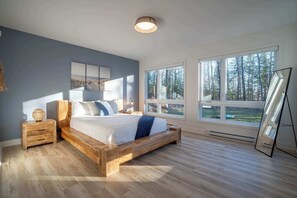 Une chambre à coucher avec un lit en bois, une table de chevet, un miroir et de grandes fenêtres donnant sur la forêt.
