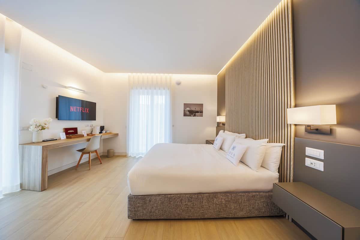A modern hotel room with a large bed, a desk with a computer, a TV mounted on the wall, and a window with curtains.