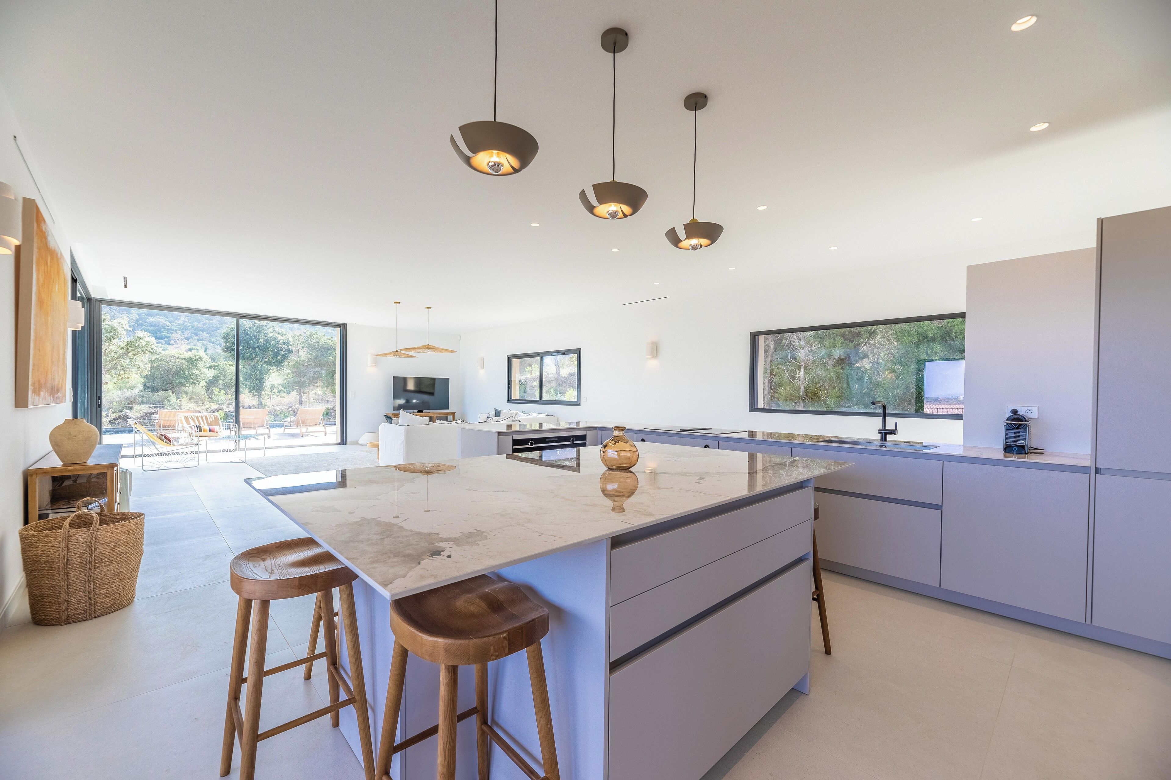 A modern kitchen with a large island, pendant lighting, and large windows offering views of the outdoors.