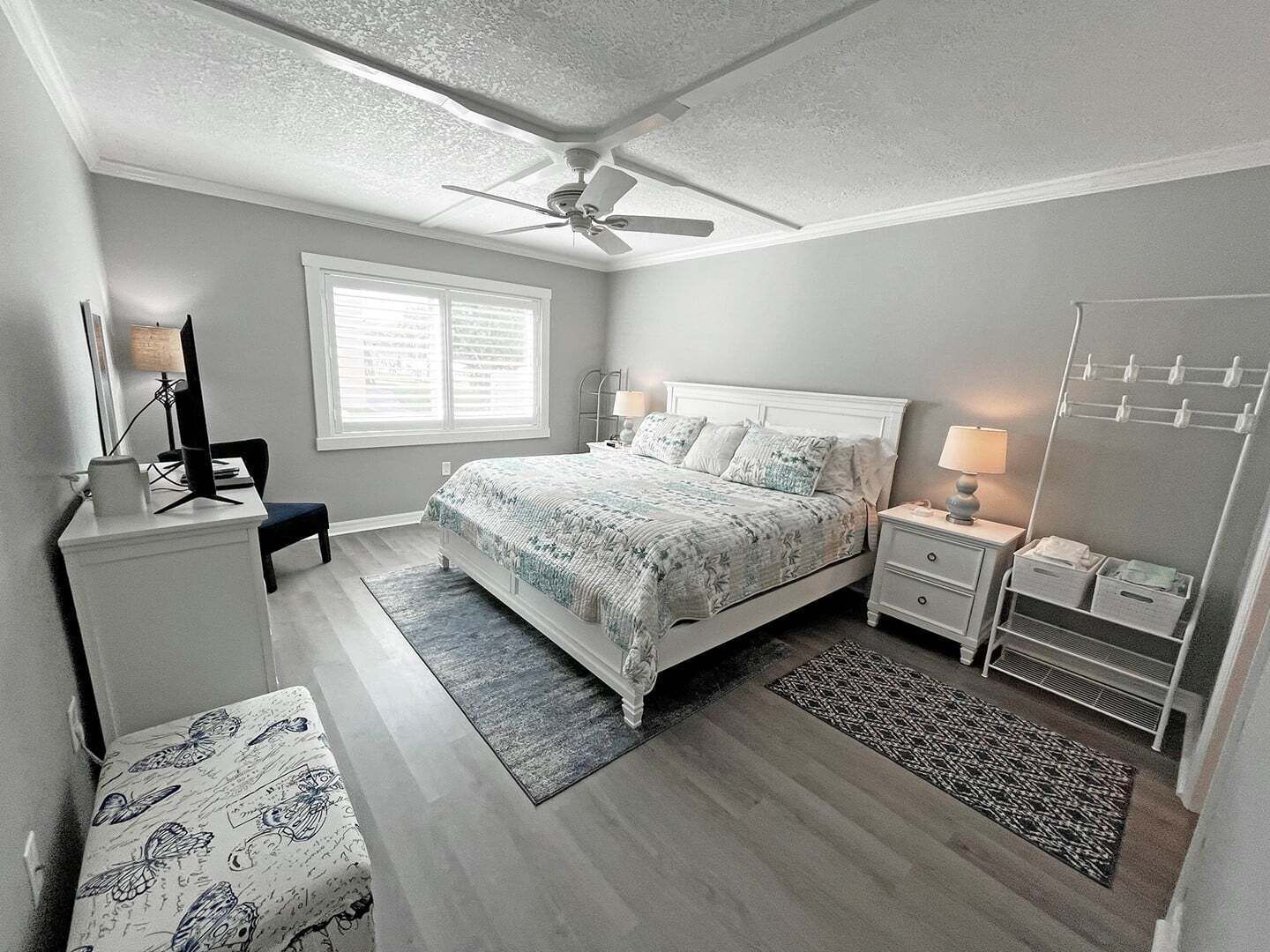 A bedroom with a bed, bedside tables, a desk, a chair, a ceiling fan, and a window with blinds.