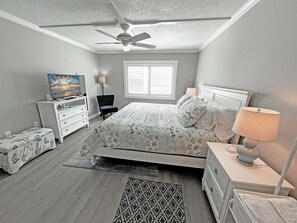 A bedroom with a bed, a nightstand, a dresser, a ceiling fan, a window with blinds, and a television on a dresser.