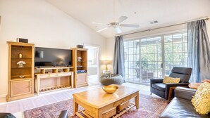 A spacious living room with a flat-screen TV, wooden entertainment unit, comfortable seating, and large windows offering a view of the outdoors. - 133 River Crossing - Barefoot Resort Rentals (North Myrtle Beach)