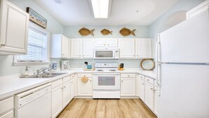 Une cuisine avec des meubles blancs, un four à micro-ondes, une cuisinière et un réfrigérateur. Trois décorations murales représentant des poissons et un panier ornent le mur.