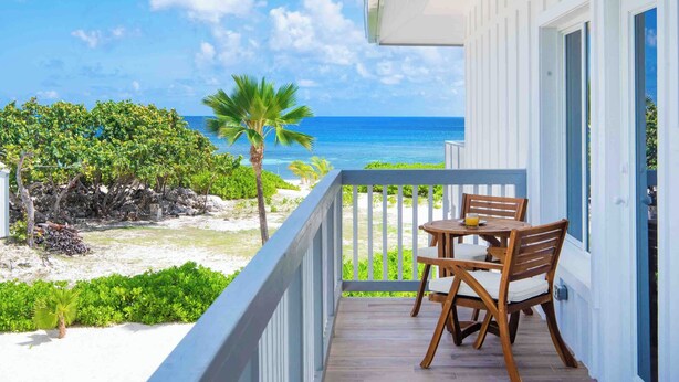 Ein Balkon mit Holztisch und -stühlen, der Blick auf einen Strand mit Palmen und dem Meer freigibt.