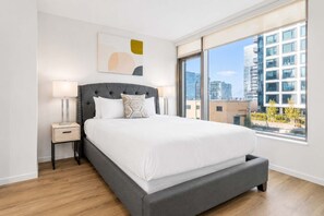 Un dormitorio moderno con una cama grande, una mesita de noche, una lámpara y vistas de la ciudad a través de amplias ventanas.