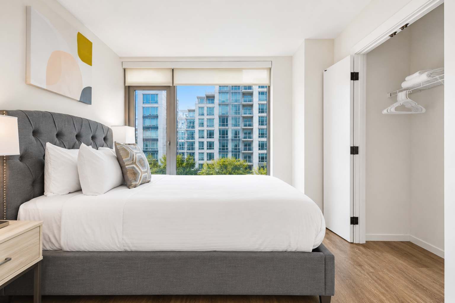 A bedroom with a large bed, a bedside table, a window with a view of buildings, and a closet with hanging clothes.