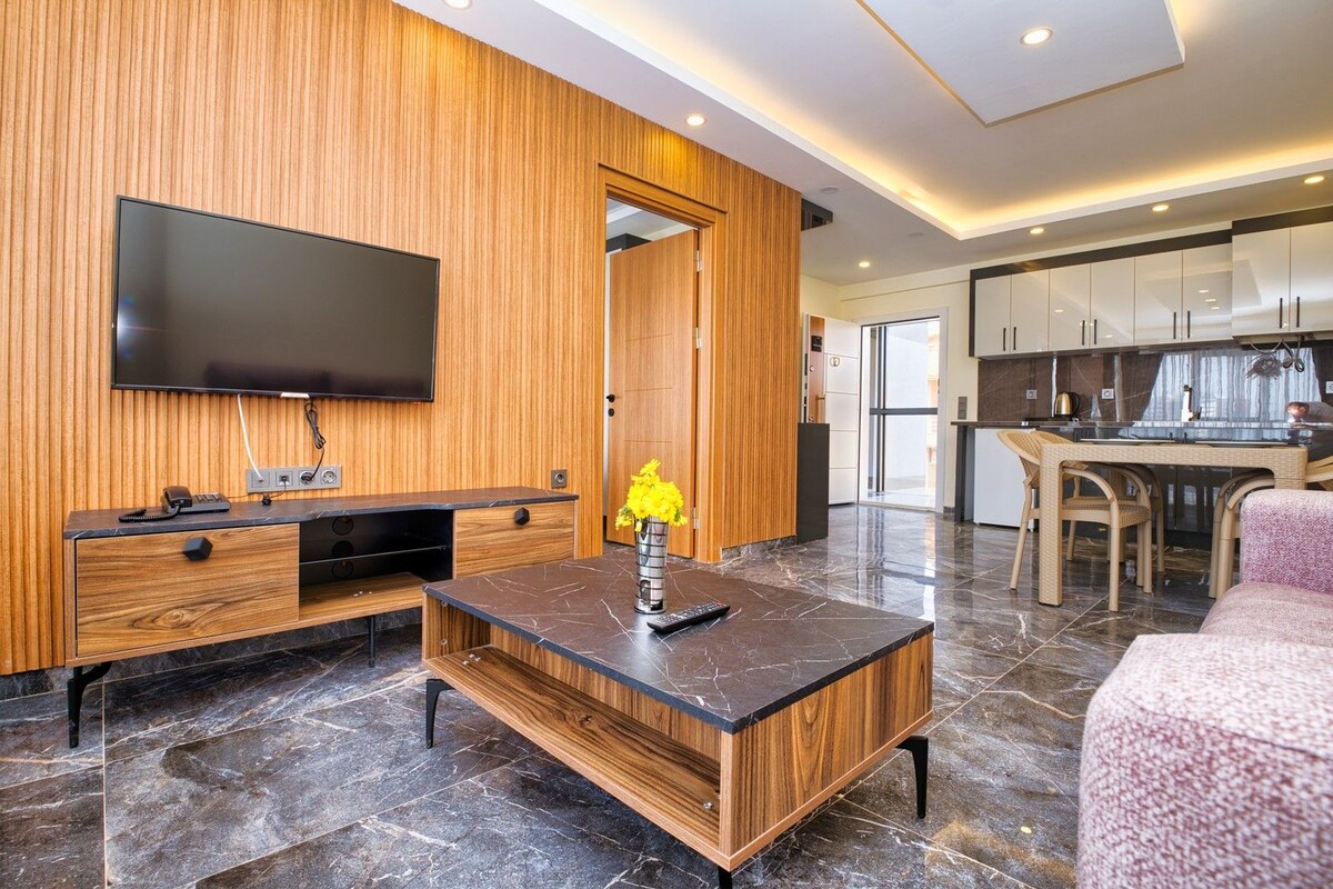 A modern living room with a flat-screen TV, wooden paneling, a marble-tiled floor, a dining area with wicker furniture, and a kitchen in the background.