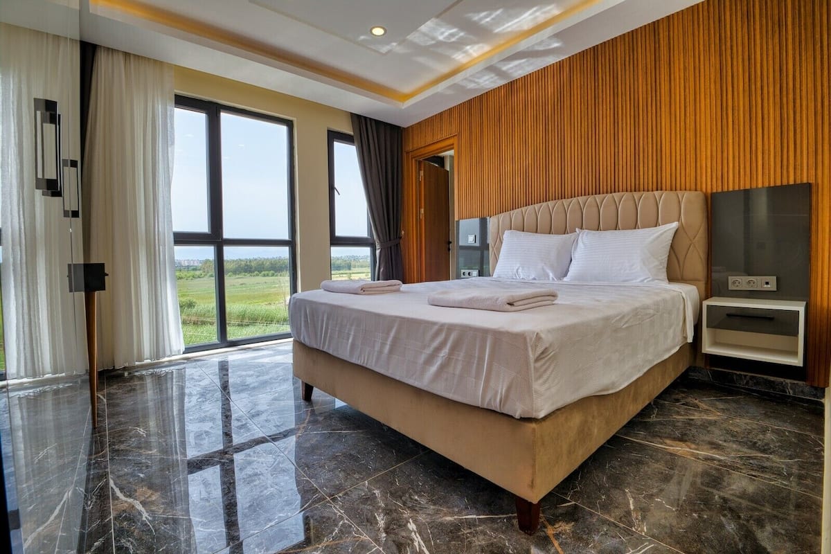 A hotel room with a large bed, a nightstand, a TV, and a view of the outdoors.