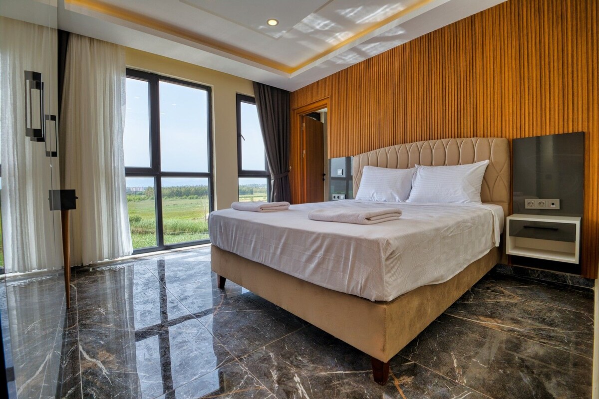 A hotel room with a large bed, a nightstand, a TV, and a view of the outdoors.
