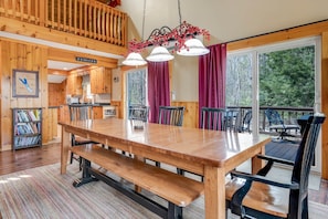 A wooden interior with a large dining table, benches, and chairs. There is a kitchen area with wooden cabinets and a hanging light fixture. - Lake Front Cabin on Desirable Highland Lake (Bridgton)
